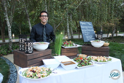 Contemporary market salad display with server ready to meet guests. Outdoor event with lanterns, grass, wood and porcelain display pieces